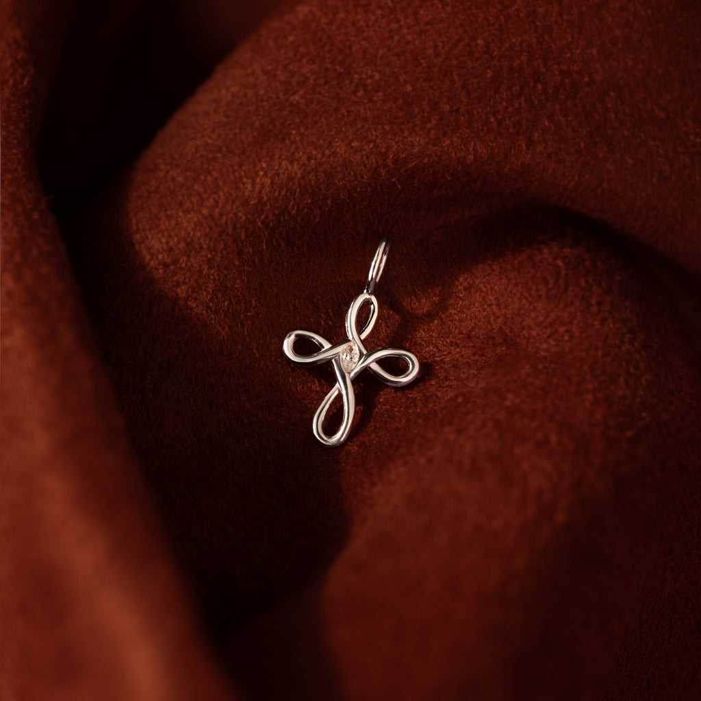 The Infinity Cross Charm in Silver rests on rich brown velvet, a meaningful everyday accessory that beautifully symbolizes eternal faith.
