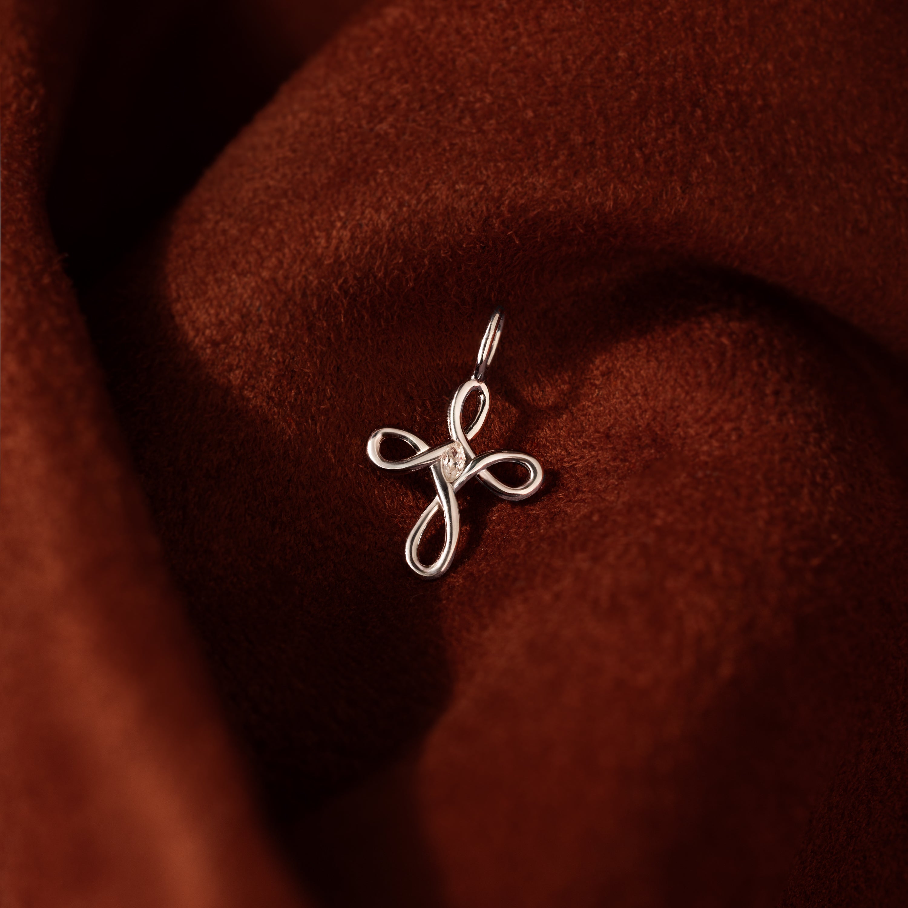 The Infinity Cross Charm in Silver rests on rich brown velvet, a meaningful everyday accessory that beautifully symbolizes eternal faith.