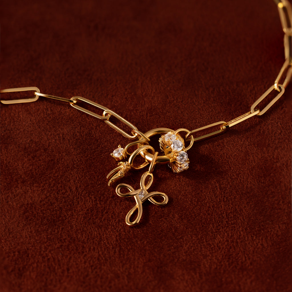 The Infinity Cross Charm in 18K Gold is shown on a brown surface, featuring exquisite jeweled accents, a floral pendant, and meaningful gold chain necklace charms.