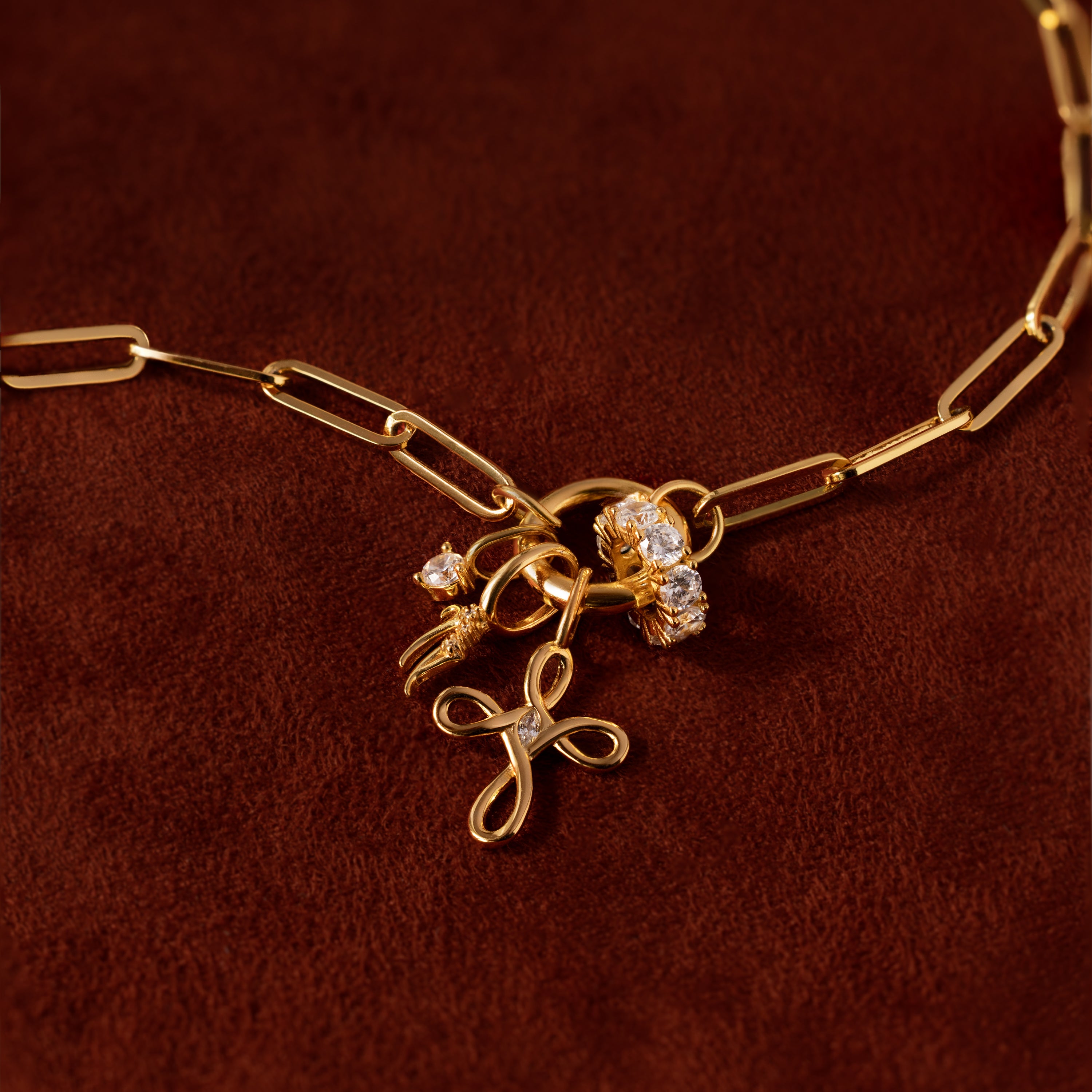 The Infinity Cross Charm in 18K Gold is shown on a brown surface, featuring exquisite jeweled accents, a floral pendant, and meaningful gold chain necklace charms.