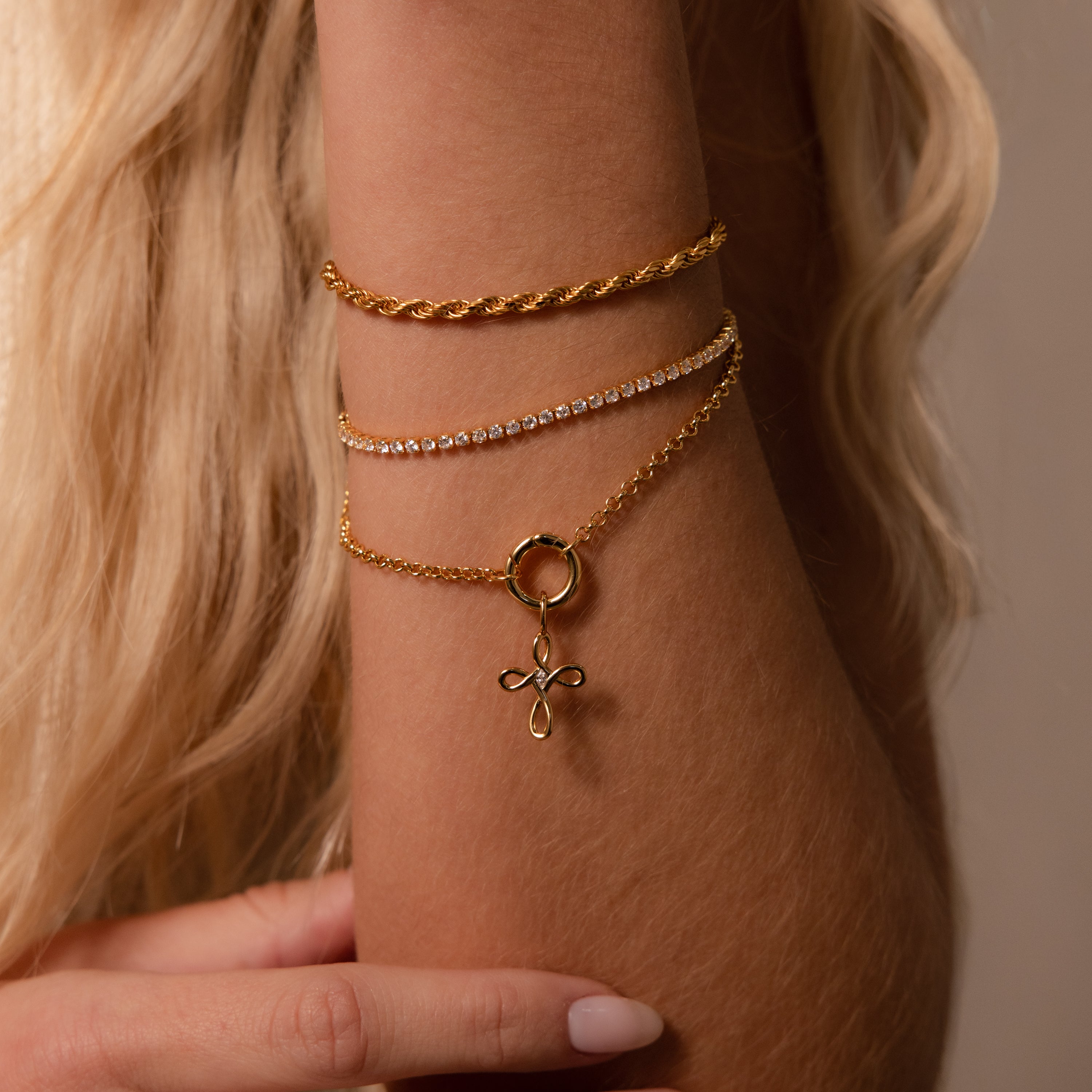 A close-up of a wrist with three gold bracelets, one adorned with the Infinity Cross Charm in Silver, adding a symbol of eternal faith, set against wavy blonde hair.