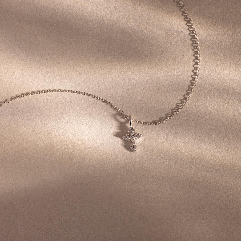 The Vintage Cross Necklace in Silver is a minimalist piece featuring a pavé-finish, diamond-studded cross pendant, shown on a beige textured surface.