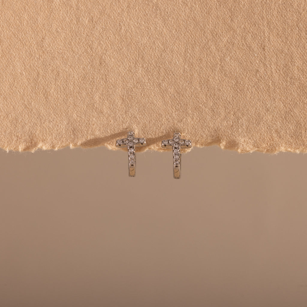 The Diamond Cross Huggies, featuring small silver crosses with pavé stones, are displayed against a textured beige background.