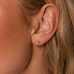 A close-up shows an ear wearing the Diamond Cross Huggies—a small gold hoop with pavé stones on a delicate cross charm, perfect for lovers of dainty, sparkling jewelry.