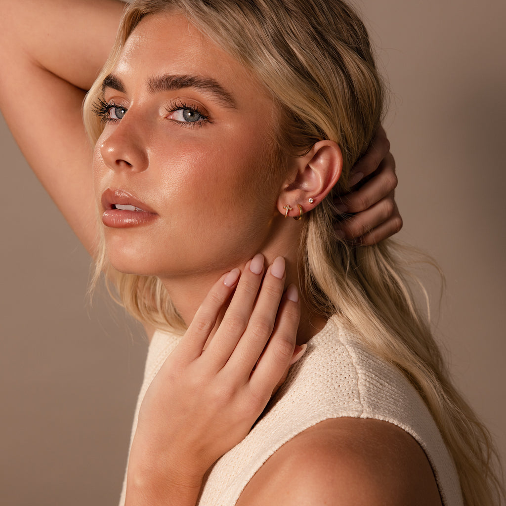 A blonde woman wearing natural makeup and a sleeveless top poses with her hand on her neck, showcasing elegant Diamond Cross Huggies.