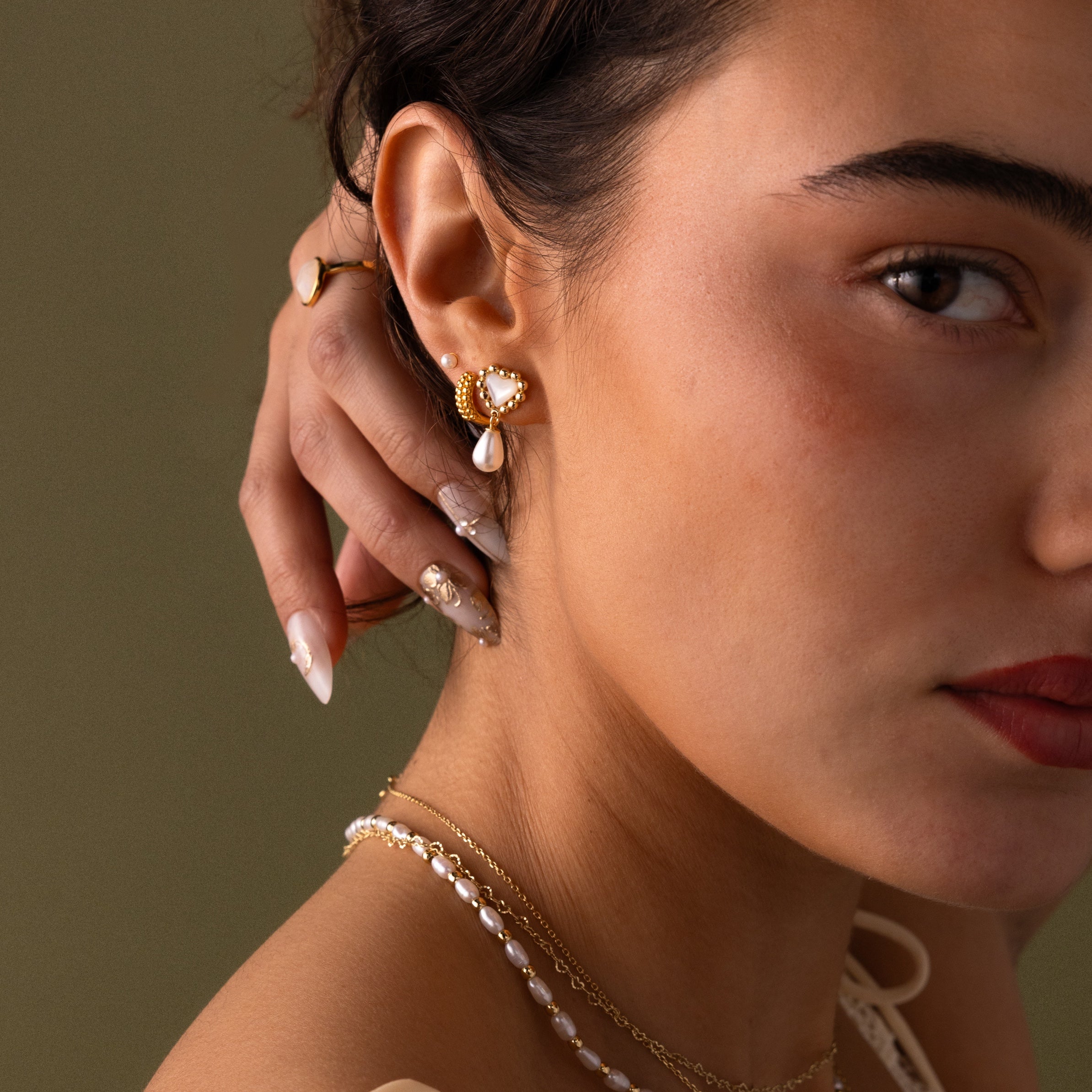 Woman touching her ear, wearing Pearl Heart Beaded Drop Studs in 18K Gold with a pearl necklace against a neutral background—an ideal bridesmaid gift.