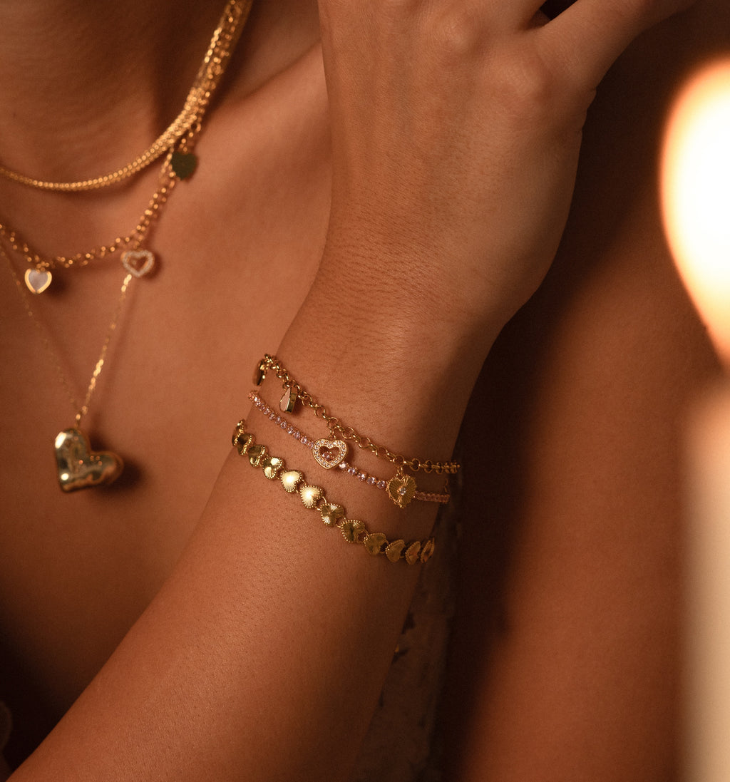 A woman wears layered gold necklaces and the Heart Charm Bracelet in 18K Gold with heart and disc charms, holding her hand near her face—a perfect gift for your girlfriend.