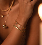 A woman wears layered gold necklaces and the Heart Charm Bracelet in 18K Gold with heart and disc charms, holding her hand near her face—a perfect gift for your girlfriend.