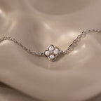 The Rounded Pearl Flower Bracelet in Sterling Silver features a four-petal floral charm, each petal adorned with a small pearl, creating an elegant accessory showcased on a beige surface.