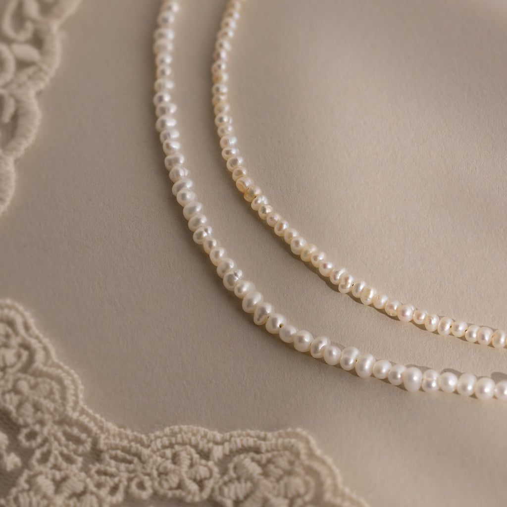 Two Summer Pearl Beaded Necklaces are displayed on beige fabric with lace corners, highlighting the timeless elegance of modern pearl jewelry.