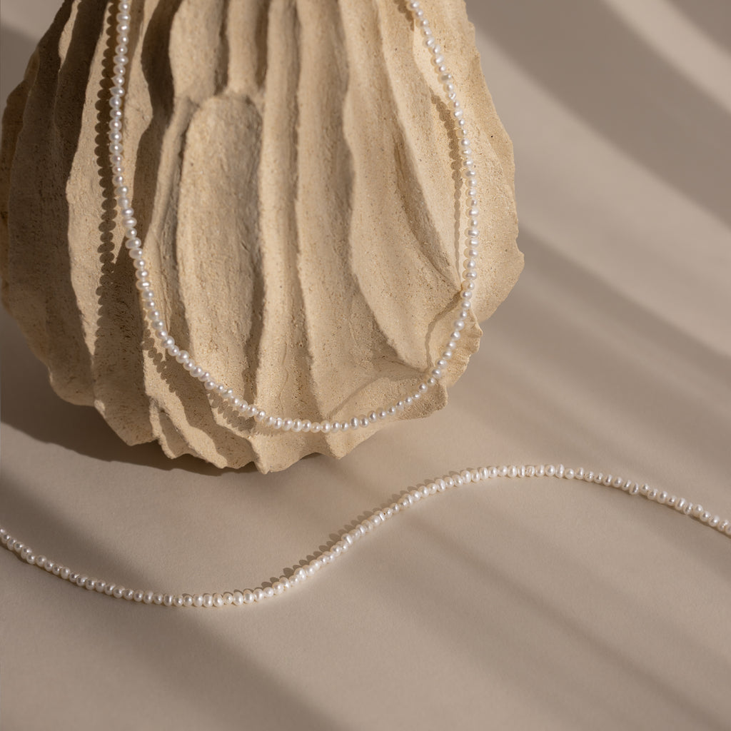 The Summer Pearl Beaded Necklace in Sterling Silver is displayed draped over a textured beige stone, with soft shadows falling across a neutral background for a modern, elegant look.