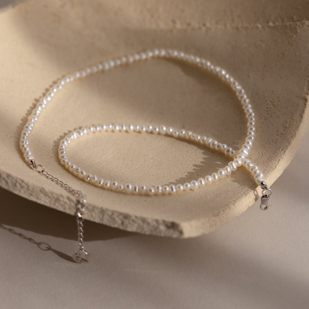 The Summer Pearl Beaded Necklace in Sterling Silver features delicate pearls and a silver clasp, offering a modern take on pearl jewelry. Shown on a textured beige surface.