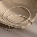 The Summer Pearl Beaded Necklace in Sterling Silver features delicate pearls and a silver clasp, offering a modern take on pearl jewelry. Shown on a textured beige surface.