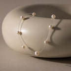 The Floating Pearl Necklace features delicate freshwater pearls on a clear strand, creating a graceful floating effect as it drapes over a smooth, round beige ceramic bowl.