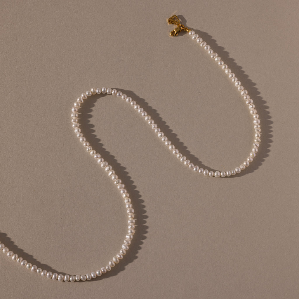 The Venus Pearl Necklace, a single-strand freshwater pearl necklace with a gold clasp, rests on a beige background, showcasing timeless elegance.