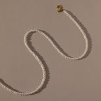 The Venus Pearl Necklace, a single-strand freshwater pearl necklace with a gold clasp, rests on a beige background, showcasing timeless elegance.