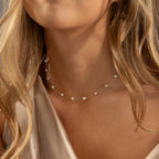 A woman with wavy blonde hair wears the Floating Pearl Necklace, featuring delicate freshwater pearls, paired with a silky beige top.