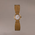 The Brick Link Timepiece is a gold sculptural watch with a round face and bold brick-link bracelet, showcased on a plain, light brown background.