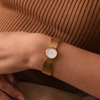 The Brick Link Timepiece, featuring a gold brick-link bracelet and white face, graces a person’s wrist against a brown ribbed top.