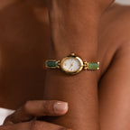 A person wears the Jade Band Timepiece, a gold wristwatch featuring a white minimalist oval dial and green jade band accents, while touching their arm.
