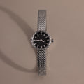 Silver braided timepiece with a black face centered on a neutral background.