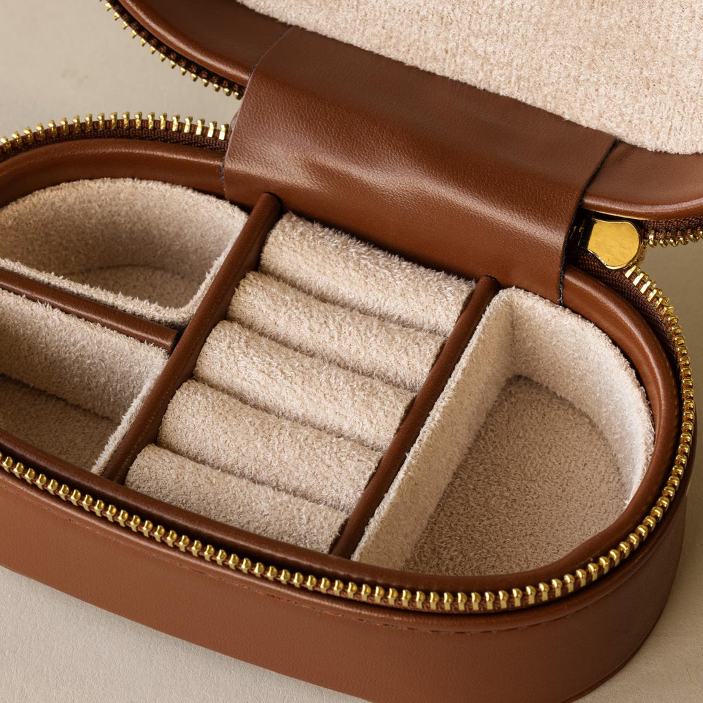 The Oval Leather Jewelry Box features a brown exterior, beige lining, ring slots, and section dividers—an ideal compact organizer to keep your jewelry neat and tidy.
