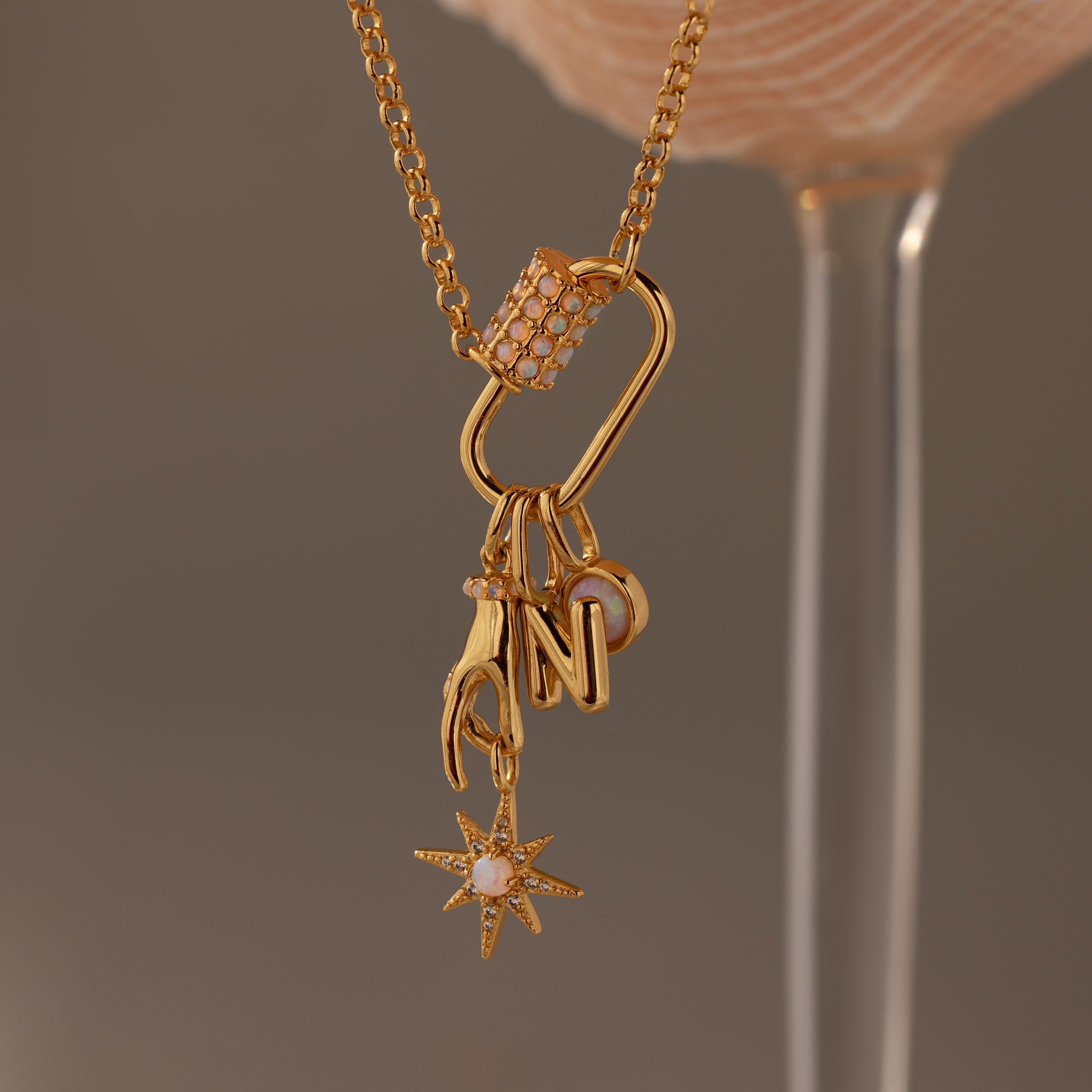 Gold necklace with hand, letter N, and star charms displayed on a soft pink background. The Opal Carabiner Clasp doubles as a luminous charm connector, adding an elegant opal accent to the design.