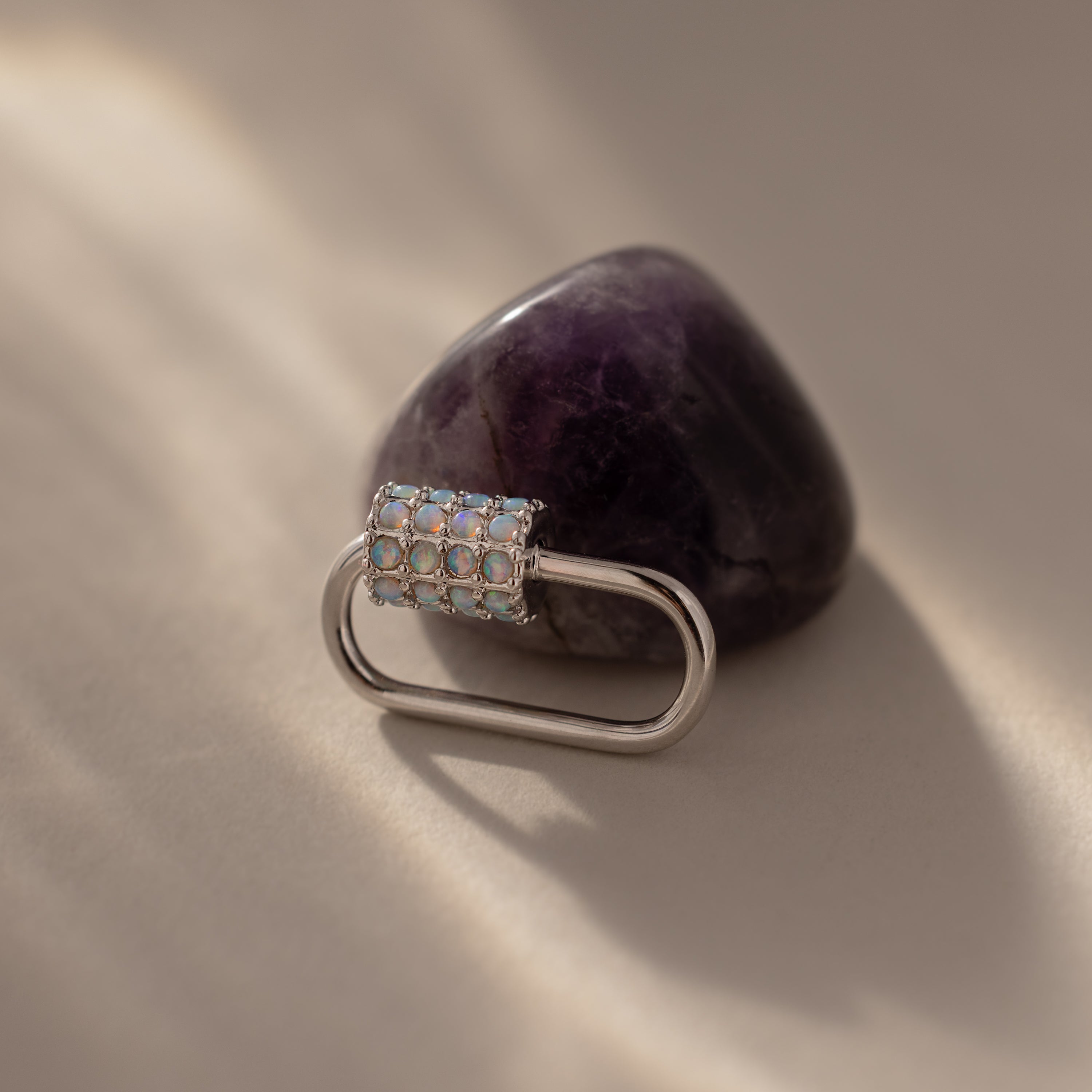 A white gold, oval-shaped Opal Carabiner Clasp with gems is displayed on a polished amethyst stone atop a beige surface.