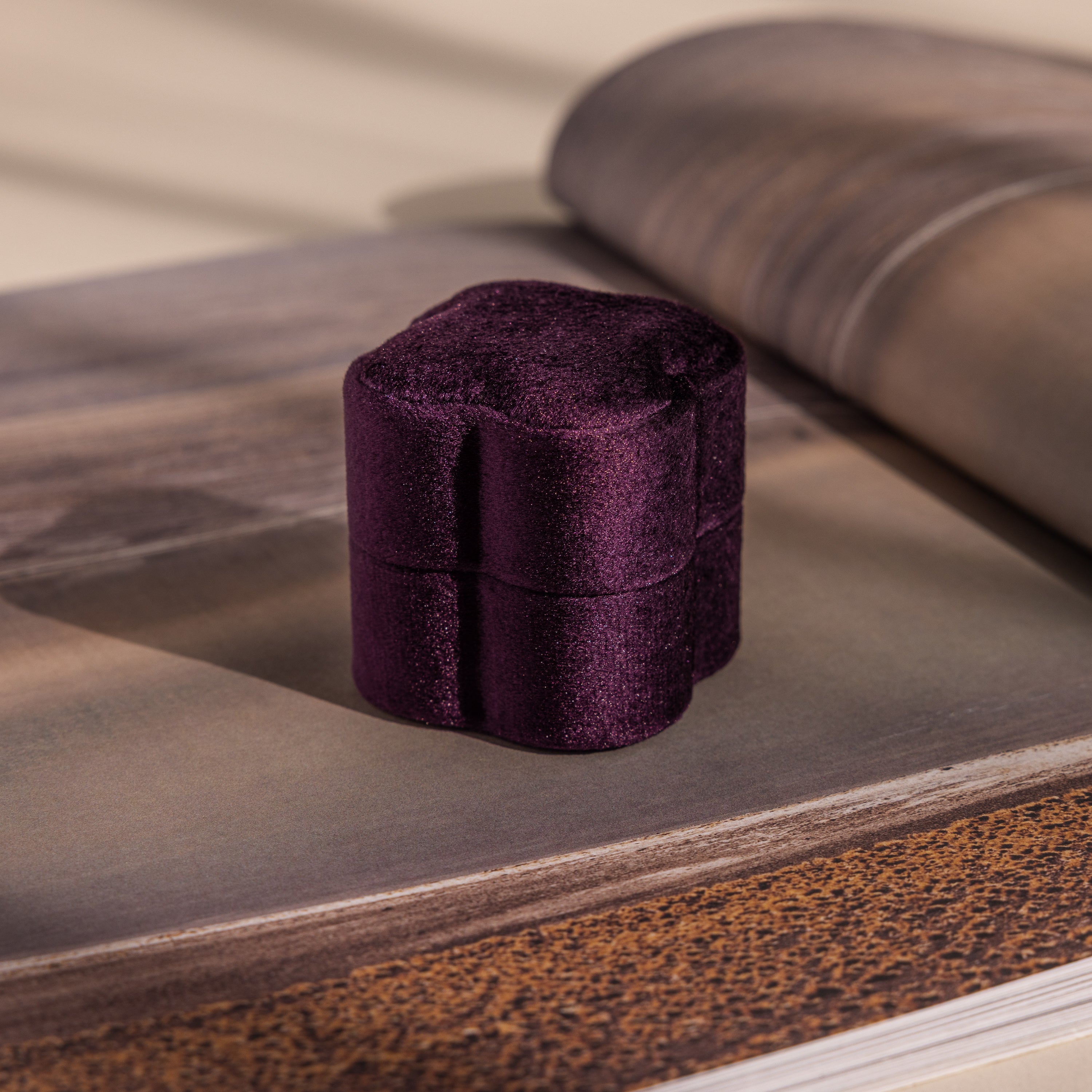 The Purple Flower Velvet Ring Box, small and elegant, rests on an open book against a softly lit, neutral background.