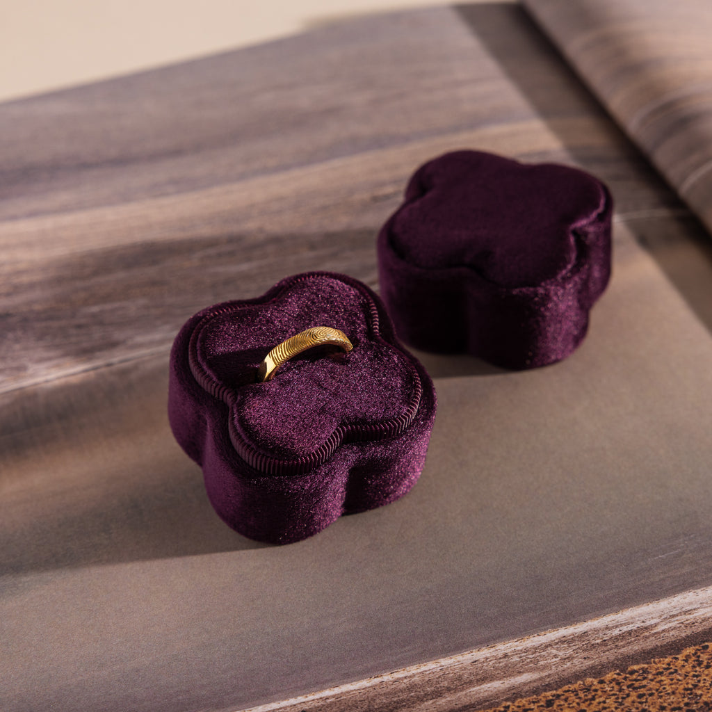 A gold ring rests in a Purple Flower Velvet Ring Box with its lid beside it on a textured surface, the box's deep purple color enhancing the elegant, dreamy scene.
