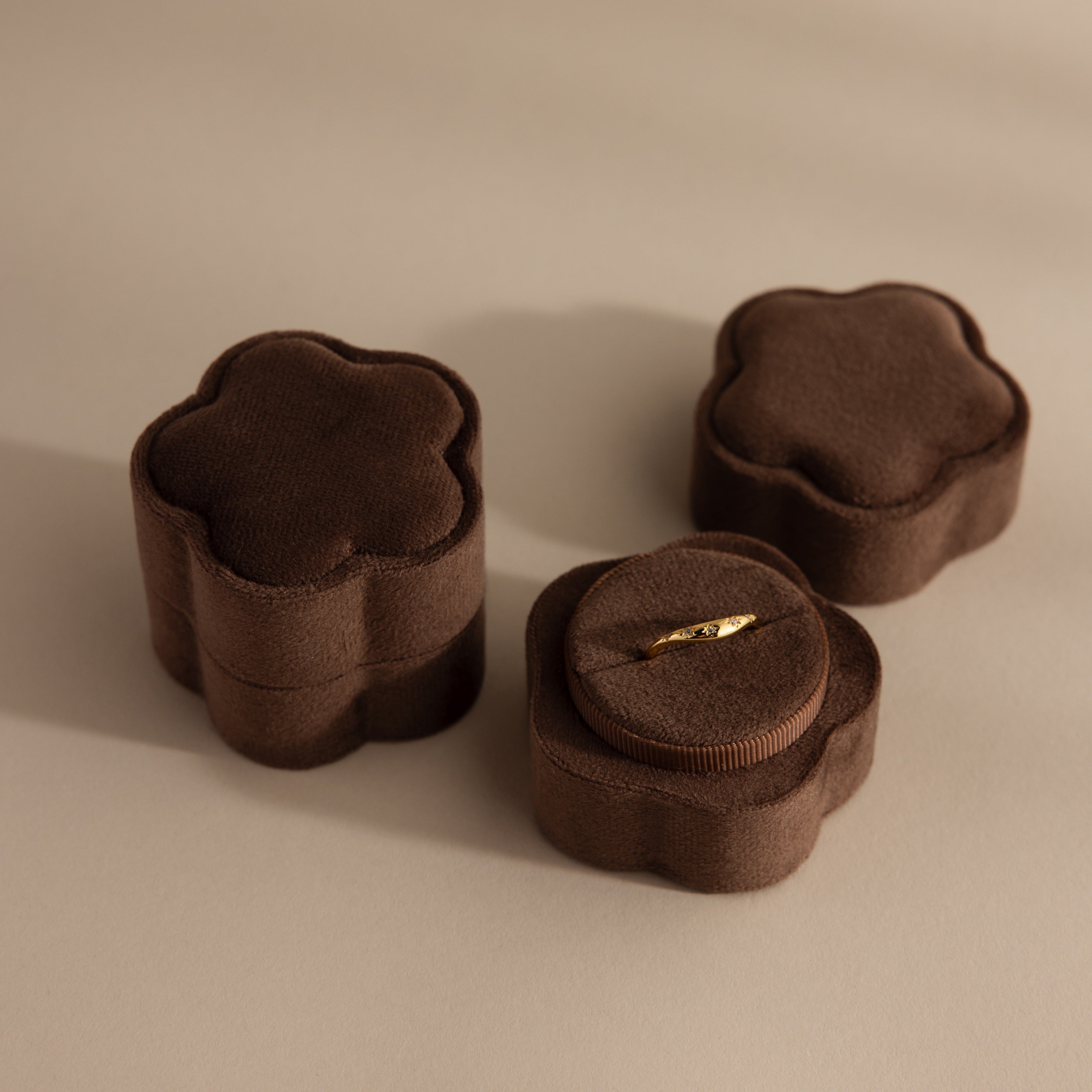 A Brown Flower Velvet Ring Box holds a delicate gold ring, with two matching closed boxes placed nearby.