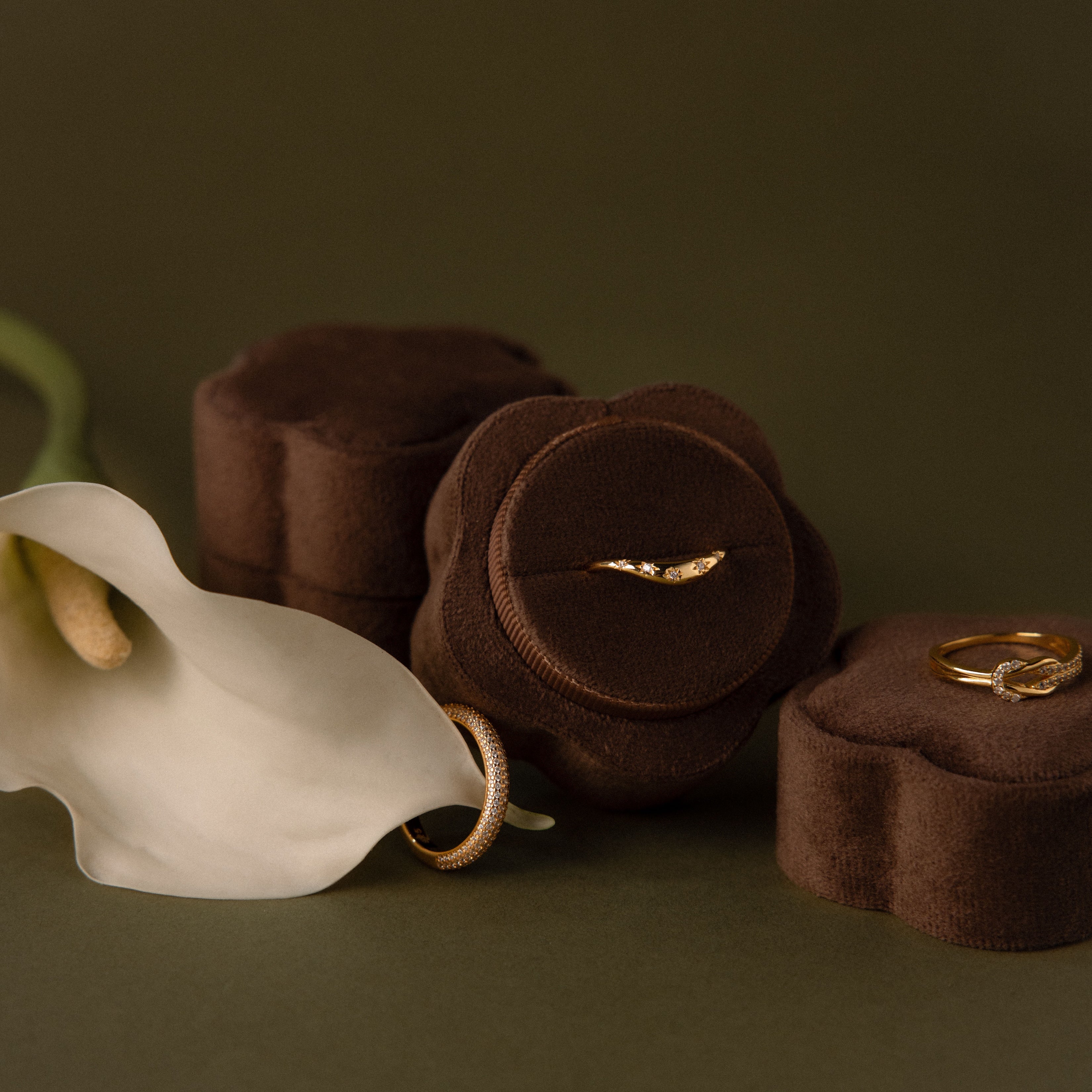 Gold rings displayed in a Brown Flower Velvet Ring Box beside a white calla lily, set against a dark background.