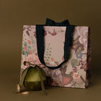 Floral gift bag with black handles, a green ornament, and two gold rings beside the Velvet Round Ring Box on an olive background.