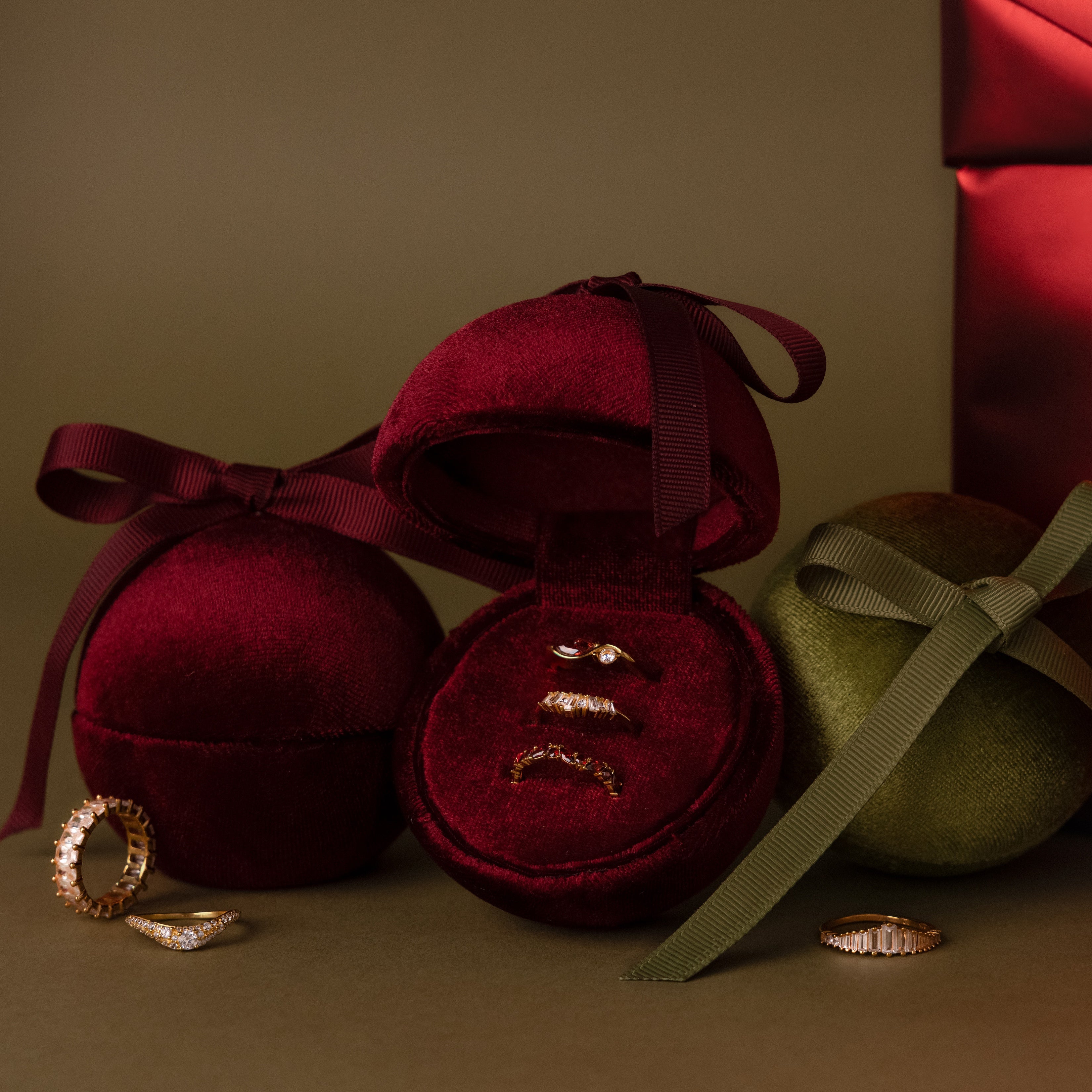 The Velvet Round Ring Box with ribbon bow holds gold rings and is displayed on a neutral background alongside a red jewelry gift box.
