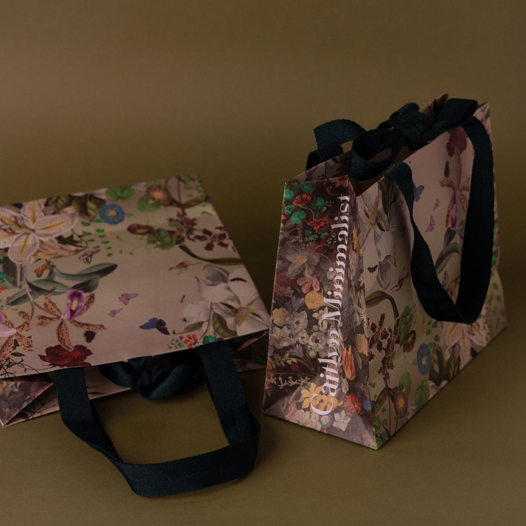 The Floral Gift Bag Add-On features two elegant floral-patterned gift bags with black ribbon handles, providing a special finishing touch—all set against a brown background.