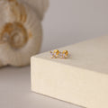 Butterfly Alexandrite Flatback Studs with small crystals, ideal as personalized jewelry, are displayed on a beige surface with a seashell in the background.