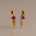 The Floating Amethyst Huggies, featuring gold earrings with a central purple gemstone, are showcased against a plain beige background.