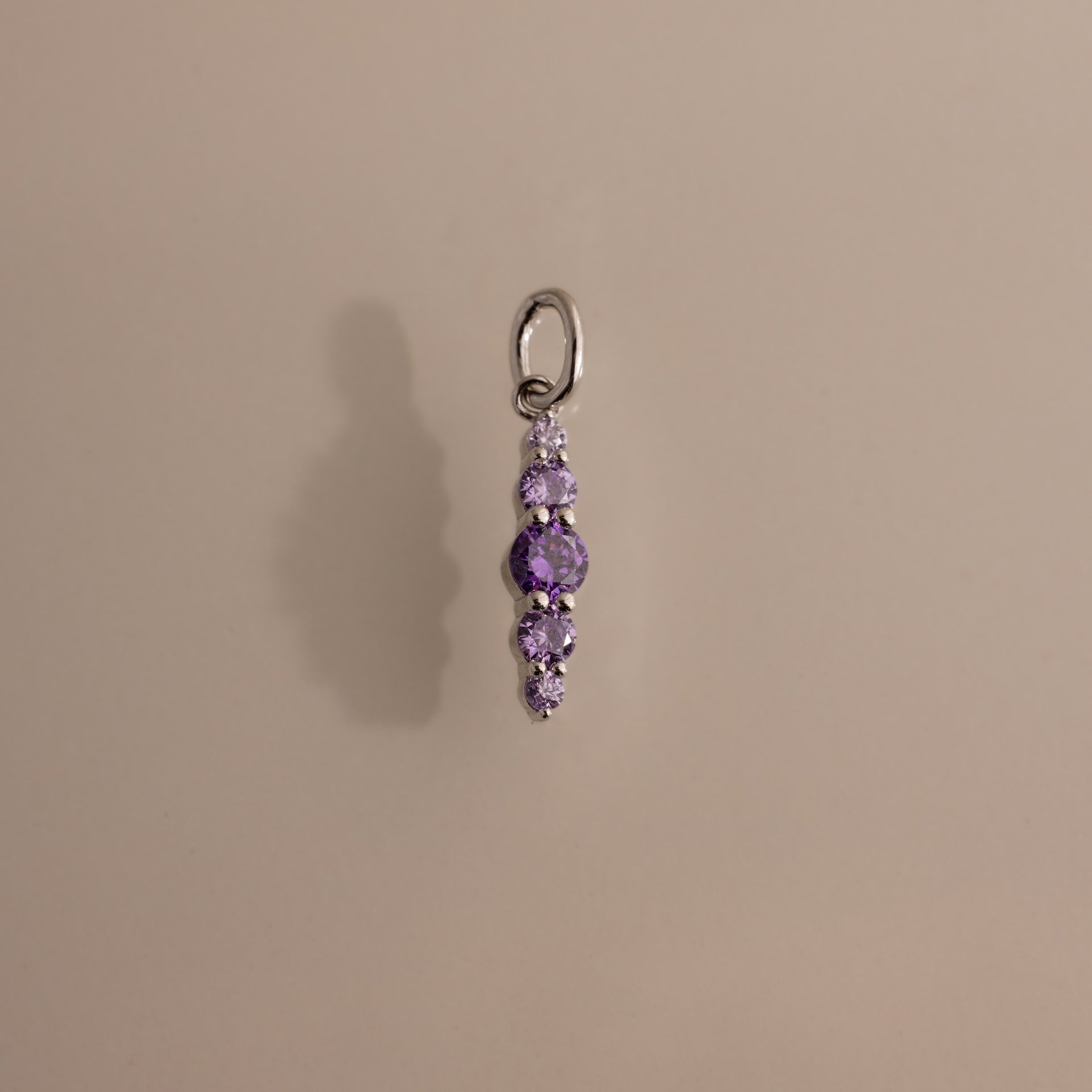 The Ombre Lace Amethyst Charm is a silver pendant with five vertically arranged purple gemstones, ideal as a personalized charm or birth month gift, shown on a light beige background.