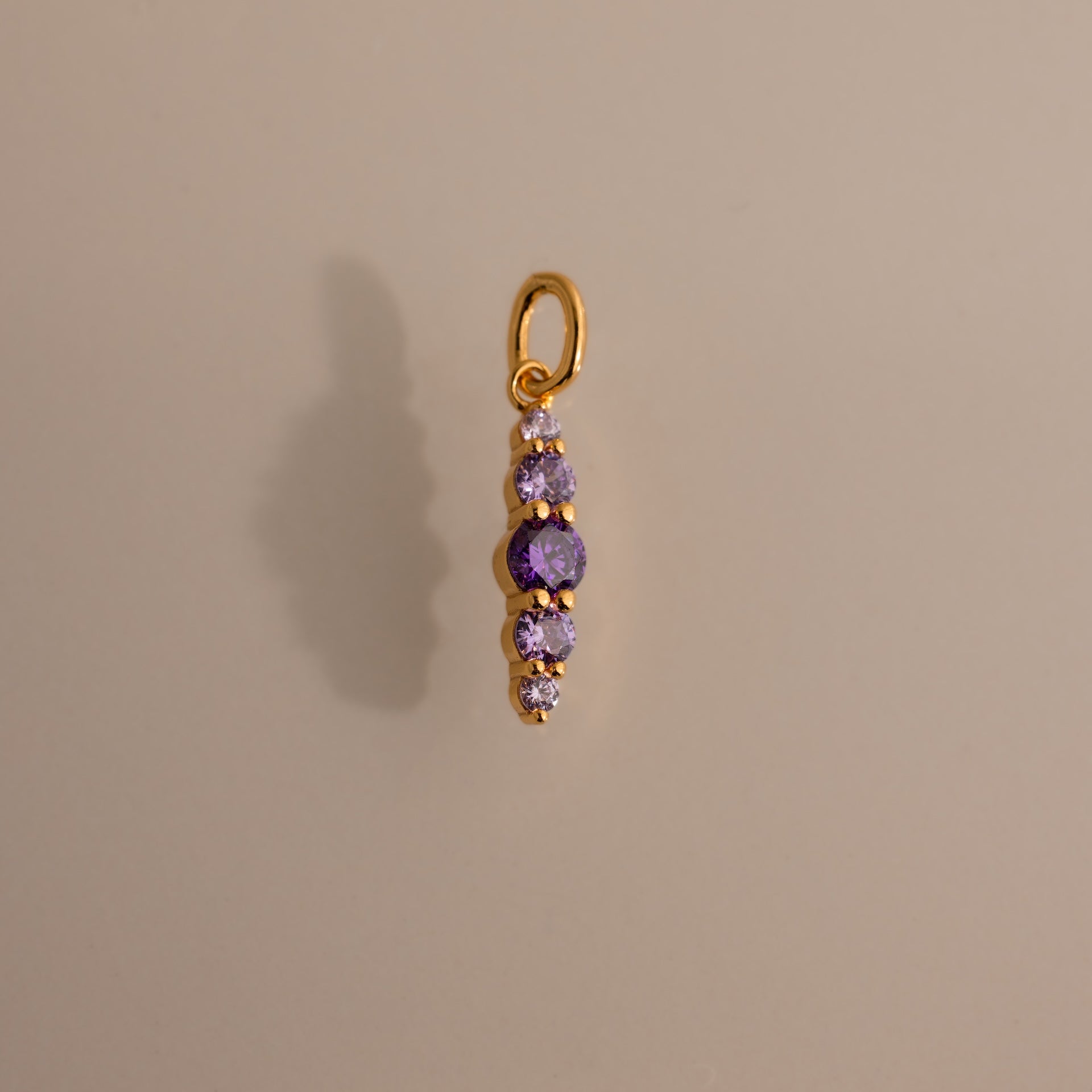 The Ombre Lace Amethyst Charm features five vertically arranged purple gemstones and casts a shadow on a beige background—an ideal personalized birth month gift.