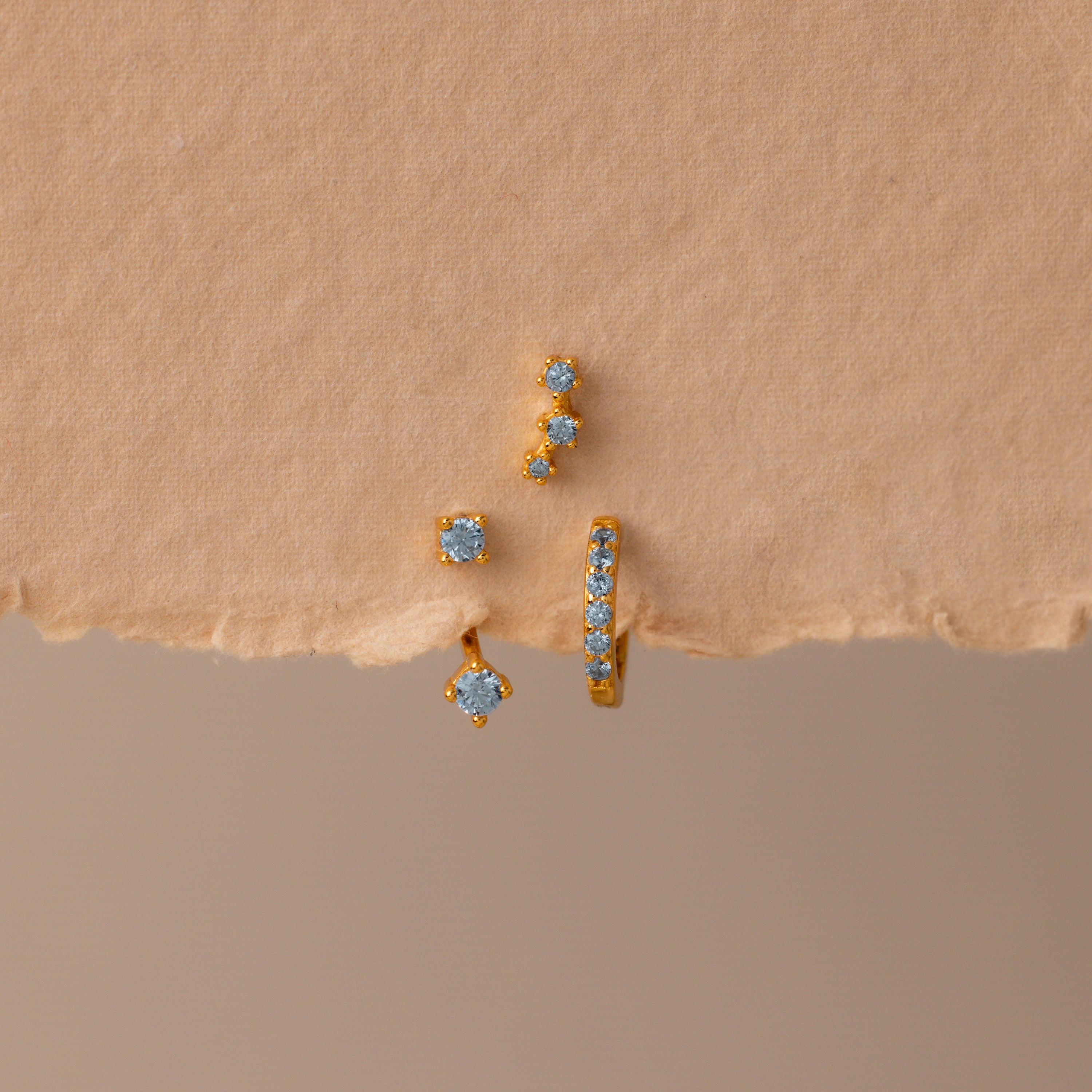 The Aquamarine Earring Trio Set features three gold earrings with blue gemstones—a delicate huggie hoop, an elegant ear climber, and a third design—displayed on textured beige paper.