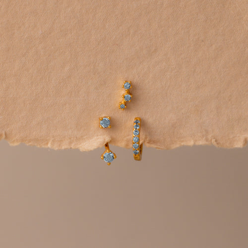 The Aquamarine Earring Trio Set features three gold earrings with blue gemstones—a delicate huggie hoop, an elegant ear climber, and a third design—displayed on textured beige paper.