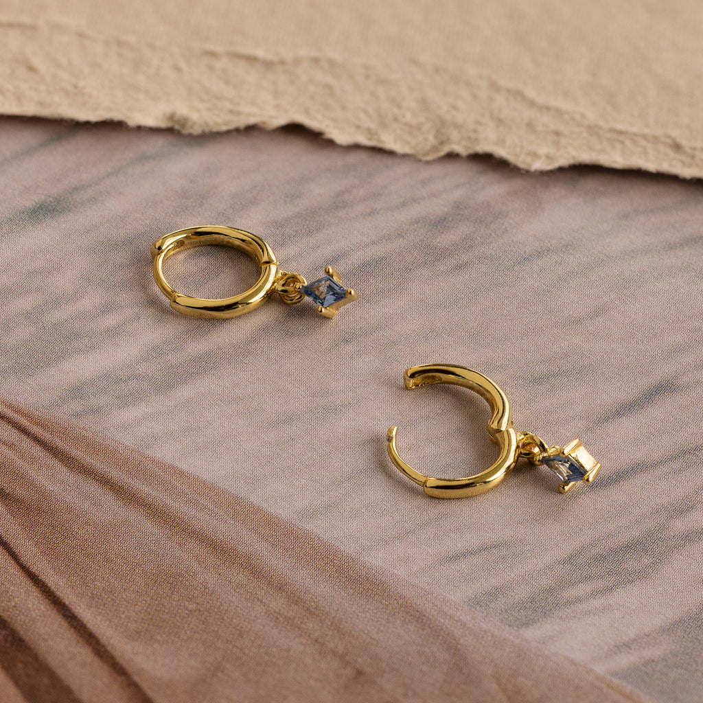 The Birthstone Drop Huggies, gold hoop earrings with square blue stones, are displayed on a beige and brown fabric surface—ideal for personalized jewelry lovers.