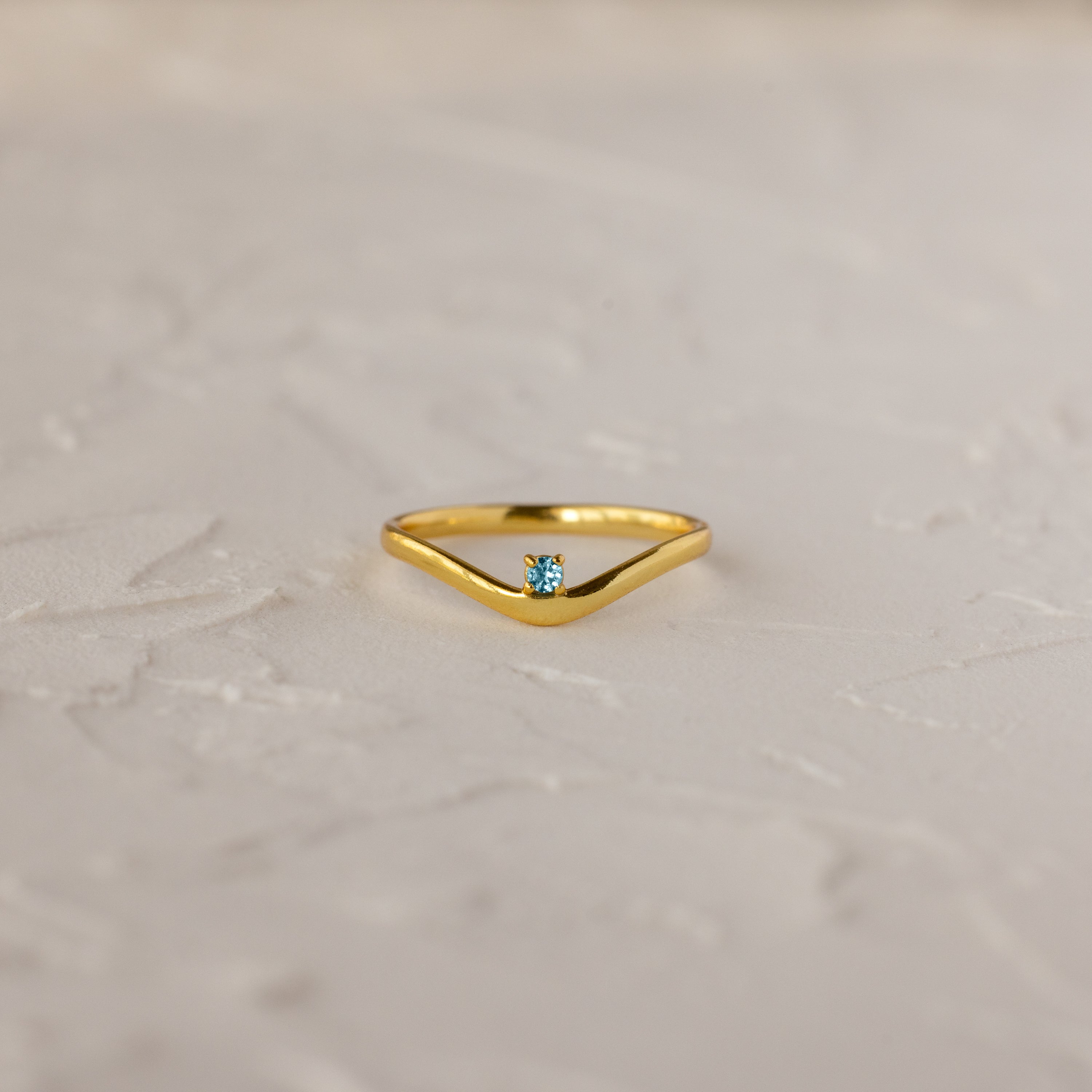The Aquamarine Drop Curve Ring, featuring a small blue gemstone, is elegantly displayed on a textured white surface.