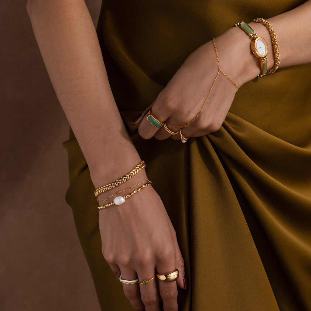 Close-up of hands adorned with gold rings, a watch, green stone jewelry, and the Pearl Charm Beaded Bracelet in 18K Gold over an olive dress.
