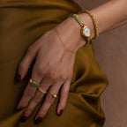 A hand adorned with the Bold Byzantine Bracelet in 18K Gold and green accents rests on golden brown satin, exuding quiet luxury.