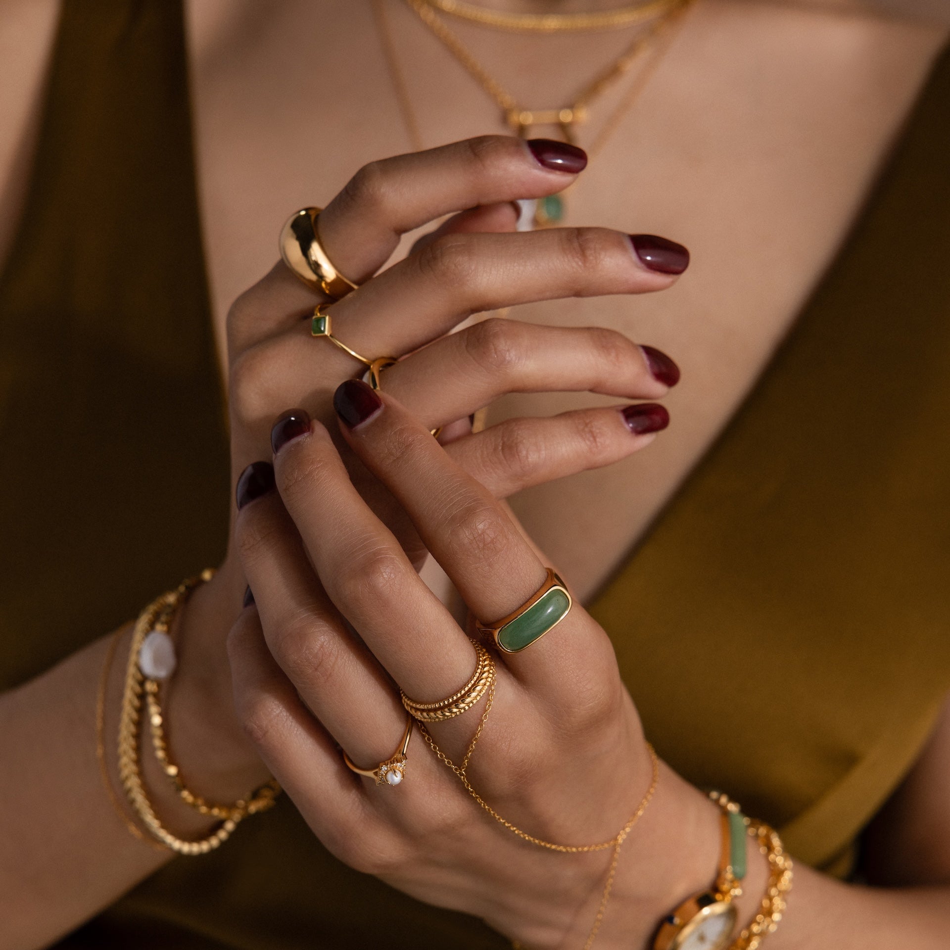 Woman with maroon nails, wearing gold jewelry and the Jade Signet Ring—a green jade ring with a gold bezel—holds her hands near her chest.