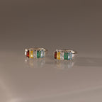 The Multiple Baguette Birthstone Huggies in Sterling Silver feature orange, yellow, clear, and green gemstones, creating a vibrant and colorful look beautifully displayed on a reflective surface.
