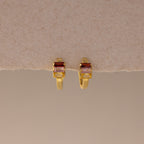 Multiple Baguette Birthstone Huggies in 18K Gold, featuring rectangular red and clear gemstones, are styled as colorful jewelry and displayed on a beige textured background.