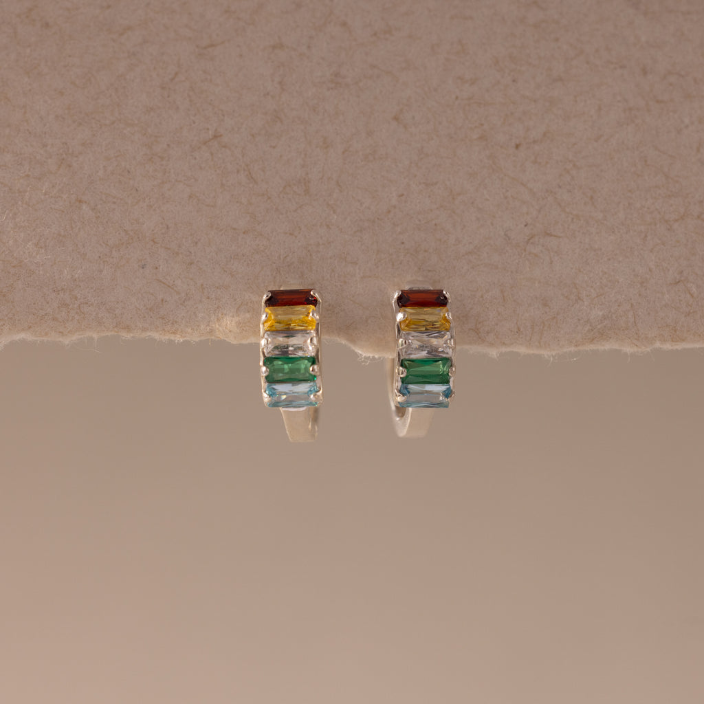 Multiple Baguette Birthstone Huggies in Sterling Silver, featuring rectangular multicolored gemstones, are beautifully showcased on a textured beige surface.