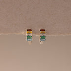 The Multiple Baguette Birthstone Huggies—silver petite hoops with rectangular red, yellow, blue, and green stones—rest on a textured beige surface, offering colorful jewelry for a playful accent to any look.